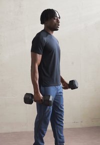 Une personne porte un t-shirt noir à manches courtes et un pantalon de sport bleu, tenant des haltères noirs, se tenant en profil devant un arrière-plan clair.