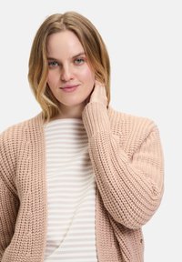 Lichtroze gebreide cardigan met een losse pasvorm, ribbelmanchetten en een gestructureerd patroon. Draag het over een wit met grijs gestreept topje.