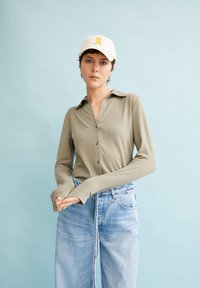 Camicia a maniche lunghe con colletto in tessuto verde oliva, abbinata a jeans chiari a vita alta. Cappello bianco con dettaglio del logo giallo.