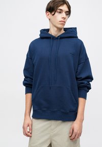 Granatowy hoodie wykonany z miękkiego materiału, z dużą kieszenią na przodzie, kapturem ze sznurkiem, ściągaczami na mankietach oraz małym logo na klatce piersiowej.
