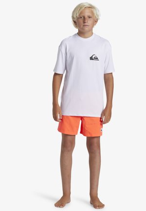 Valkoinen lyhythihainen t-paita mustalla logopocketilla, yhdistetty kirkkaan oransseihin uimapantsareihin. Kevyt kangas, yksinkertainen muotoilu ja ilman koristeluja.