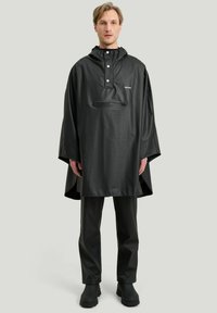 Svart vattentät poncho med huva, tryckknappar och en framficka med dragkedja. Bärs över matchande svarta byxor och stövlar. Slät textur.