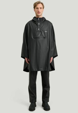 Black waterproof poncho with a hood, snap fasteners, and a front zip pocket. Worn over matching black pants and boots. Smooth texture.