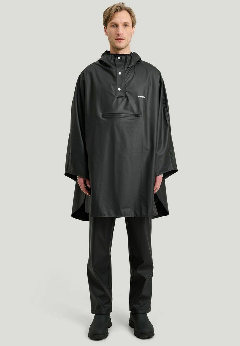 Svart vattentät poncho med huva, tryckknappar och en framficka med dragkedja. Bärs över matchande svarta byxor och stövlar. Slät textur.