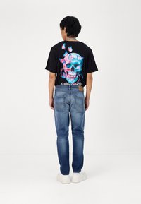 Svart t-skjorte med en levende hodeskalle-grafikk med blå og rosa detaljer, kombinert med bl�å denimjeans og hvite sneakers.