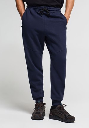 Uomo che indossa pantaloni della tuta blu scuro con tasche laterali con cerniera e scarpe da ginnastica nere, in piedi davanti a uno sfondo semplice.
