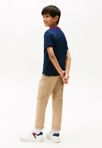 Marine polo shirt med rødt krave detalje, beige cargo bukser med sidelommer og hvide sneakers. Modellen vender væk, hænderne lagt sammen bagpå.