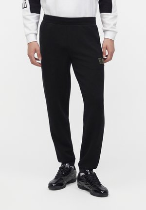 Homme portant un pantalon de jogging noir avec un logo sur la cuisse et des baskets noires brillantes, présenté de la taille aux pieds sur fond blanc.