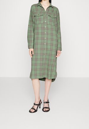 Kobieta ubrana w zieloną sukienkę z długim rękawem w kratę, zapinaną na guziki, w połączeniu z czarnymi sandałami na wysokim obcasie z paskami, stojąca na gładkiej podłodze.