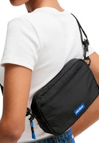 Bolso cruzado negro con un acabado texturizado, cierre de cremallera y logotipo de la marca azul. Correa ajustable y detalle de herrajes visibles.