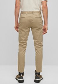 Beige slim-fit chinos med en slät textur, utrustade med två bakfickor och en bältesöggla. Kombineras med mörka, vadderade skor.