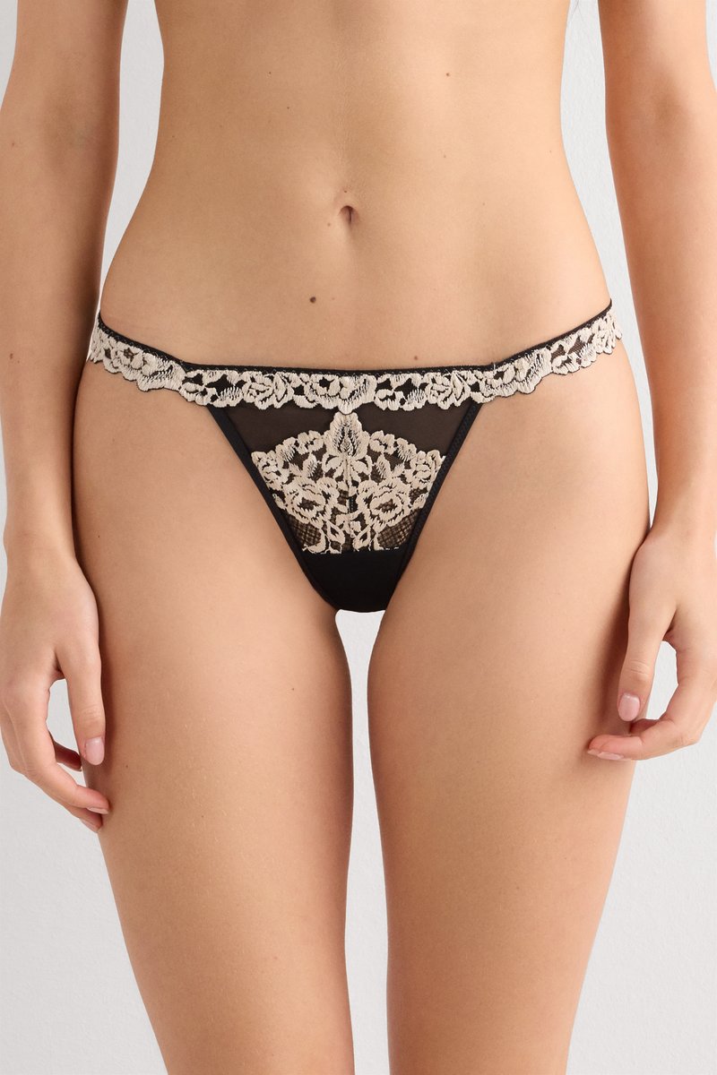 Culotte en dentelle noire transparente avec broderie florale beige portée par une personne, vue de la taille jusqu'au haut des cuisses.