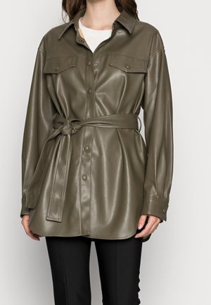 Frau trägt eine olivgrüne, kunstlederne Hemdjacke mit Gürtel, Brusttaschen mit Knöpfen und schwarze Hose, steht vor einem weißen Hintergrund.