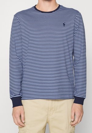 Mężczyzna ubrany w granatowo-jasnoniebieską koszulę w poziome paski z długim rękawem oraz beżowe spodnie, ręce swobodnie opuszczone wzdłuż ciała, twarz niewidoczna.