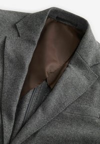 Blazer en laine grise avec un extérieur texturé. L'intérieur présente une doublure brune lisse, des coutures visibles et un éclat subtil.