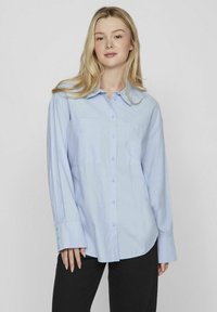 Camisa de botones azul claro con cuello, dos bolsillos frontales, mangas largas y un corte relajado. Tejido suave con una textura blanda.