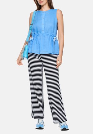 Frau trägt ein ärmelloses blaues, geknöpftes Oberteil mit seitlichen Bändern, schwarz-weiß gestreifte, weit geschnittene Hosen, blau-weiße Turnschuhe und hält eine blaue Handtasche.