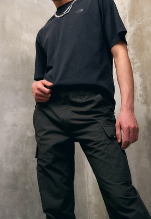 Black short-sleeve T-shirt with a subtle logo; dark green cargo pants with pockets; smooth fabric and relaxed fit. Chain accessory visible.