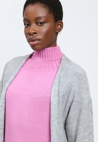 Cardigan gris claro con sutil detalle brillante, llevado sobre un cuello alto de canalé rosa. Presenta un corte holgado y una textura suave.