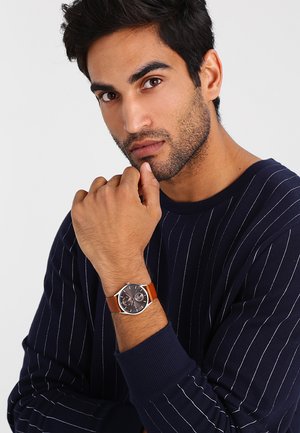 Homme aux cheveux courts et foncés avec une barbe de quelques jours, portant un pull bleu marine à fines rayures et une montre-bracelet avec un bracelet en cuir marron à son poignet gauche.