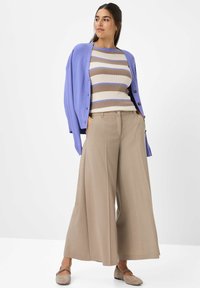 Donna in piedi con le mani nelle tasche, che indossa pantaloni beige a gamba larga, una maglietta a righe, un cardigan viola sulle spalle e scarpe flat beige.