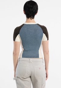 Maglietta a maniche corte con un design aderente, presenta un pannello posteriore in denim blu, spalle a contrasto marroni e corpo beige. Indossata con pantaloni grigi chiari.