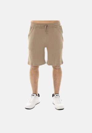 Beige geribde shorts met trekkoord gedragen met witte sneakers bij een staand persoon tegen een effen achtergrond.