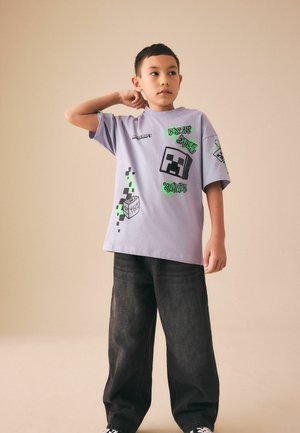 Dreng iført en lilla Minecraft-tema T-shirt og løse sorte jeans, stående med den ene hånd bag nakken mod en beige baggrund.