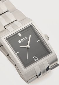 Ορθογώνιο ασημένιο μεταλλικό ρολόι χειρός με μαύρο καντράν, λευκό λογότυπο BOSS, ασημένιους δείκτες ώρας, τρεις δείκτες και ένδειξη ημερομηνίας στη θέση των 6.