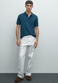 Mladý muž v modrozeleném polo tričku, bílých kalhotách a hnědých mokasínech stojící proti jednoduché světle šedé zdi.