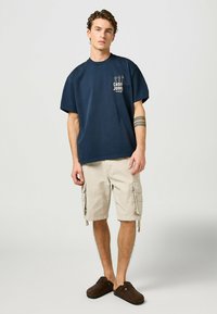 Camiseta de manga corta azul marino con un gráfico en el pecho, combinada con pantalones cortos de carga color caqui con bolsillos laterales y zuecos marrones.