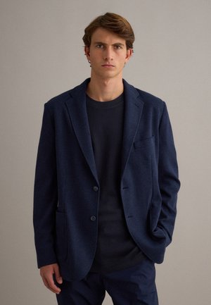 Jonge man met bruin haar, gekleed in een donkerblauw colbert over een zwart shirt, staand tegen een effen lichtgrijze achtergrond.