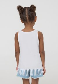 Canottiera bianca con dettagli a frange sull'orlo, abbinata a pantaloni shorts in denim blu chiaro. Il tessuto appare morbido e leggero.