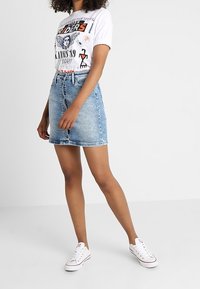 Žena v bílém tričku s potiskem, modré džínové sukni a bílých teniskách stojí před jednobarevným pozadím.