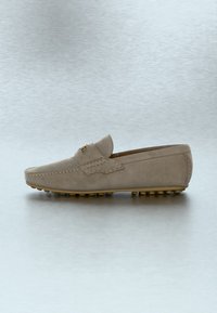 Mocassin pour homme en daim beige avec bout mocassin, coutures décoratives et semelle en caoutchouc cloutée sur un fond clair uni.