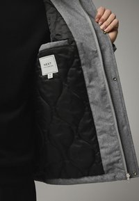 Jachetă gri cu fermoar frontal, interior negru matlasat și un buzunar interior etichetat "NEXT Tailoring" în dimensiune standard.