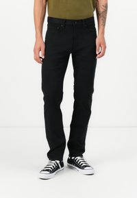 Hombre vistiendo jeans negros ajustados, zapatillas en blanco y negro, y una camisa verde oliva, de pie contra un fondo blanco.