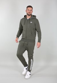 Haut à capuche vert olive avec fermeture éclair à l'avant, capuche avec cordon de serrage et poche kangourou, associé à un pantalon de survêtement assorti avec impression du logo blanc.
