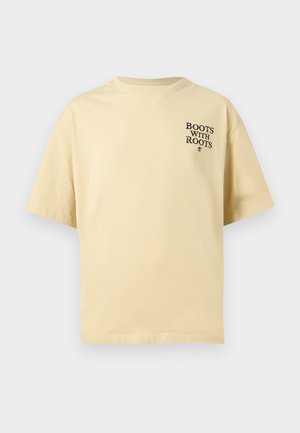 Μπεζ κοντομάνικη μπλούζα με το κείμενο «BOOTS WITH ROOTS» τυπωμένο με μαύρα γράμματα στο πάνω αριστερό μέρος του στήθους.