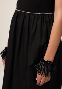 Schwarzes Kleid mit einem gerafften Rock und silberner Perlenverzierung an der Taille, kombiniert mit einem strukturierten schwarzen Fransenarmband, das lange mit Perlen besetzte Stränge aufweist.