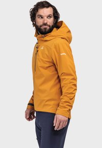 Orangene wasserdichte Jacke mit Kapuze, Reißverschluss auf der Vorderseite, verstellbaren Manschetten und Logodetail am Ärmel; kombiniert mit dunklen Hosen.