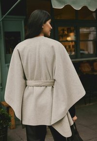 Heleroheline mähkcoat vööga, laia varrukaga ja tekstuurse kangaga. Kantud tumedate pükste peal, voolujooneline siluett.