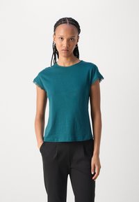 Teal kortermet t-skjorte i myk bomull med rund hals og blonde detaljer langs ermene. Kombinert med sorte bukser.