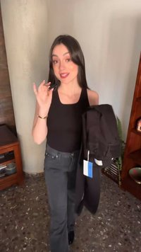 Joven sosteniendo una chaqueta negra con una etiqueta en una mano y un objeto pequeño en la otra, de pie en interiores con una sonrisa.