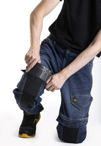 Pantalons de travail en denim bleu avec des genouillères noires, des coutures renforcées et des sangles ajustables, associés à des chaussures de sécurité noires.