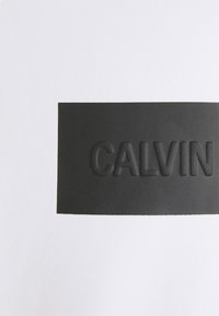 Czarna prostokątna naszywka z wytłoczonym napisem „CALVIN” na białym tle z tkaniny.