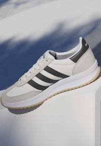Biało-szary sneaker Adidas z czarnymi paskami i gumową podeszwą, prezentowany na miękkim niebiesko-białym tle.