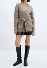 Cardigan côtelé beige avec col en V, fermeture par boutons, deux poches avant et une ceinture à nouer. Associé à une mini-jupe noire et des bottes jusqu'au genou.