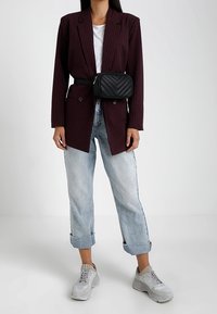 Nainen, joka pukeutunut burgundinpunainen ruudullinen bleiseri, valkoinen t-paita, käännetyt vaaleansiniset farkut, valkoiset lenkkitossut ja musta tikattu vyölaukku.