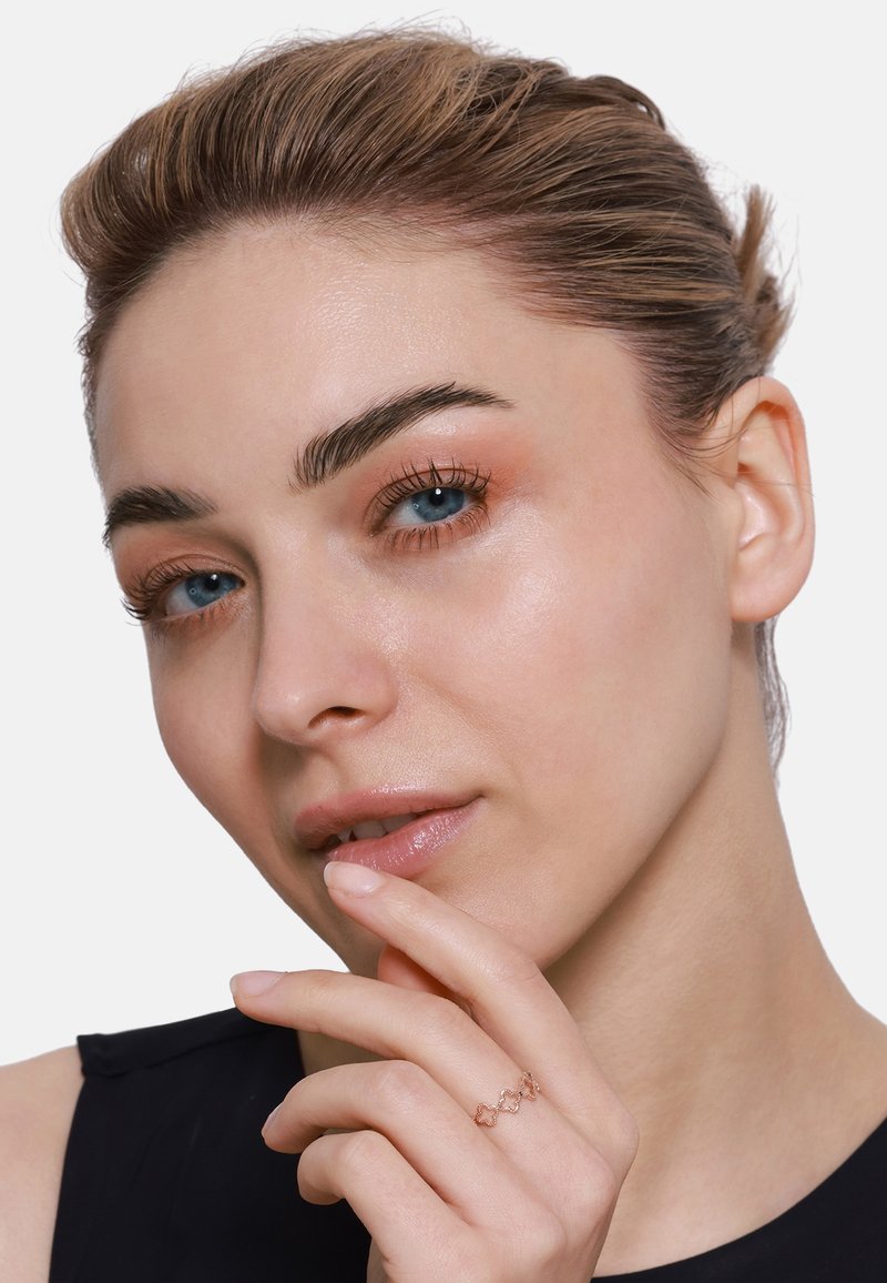Roségoldring mit geometrischem Design, der am Finger getragen wird. Das Modell hat eine klare Haut, blaue Augen und ein dezentes Make-up, vor einem neutralen Hintergrund.